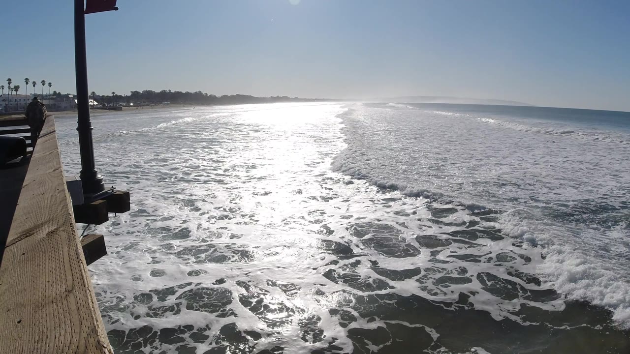 A curved line of breaking surf cuts through the sunlit water at Pismo Beach, with the pier edge on the left and open blue ocean to the right, no visible people.