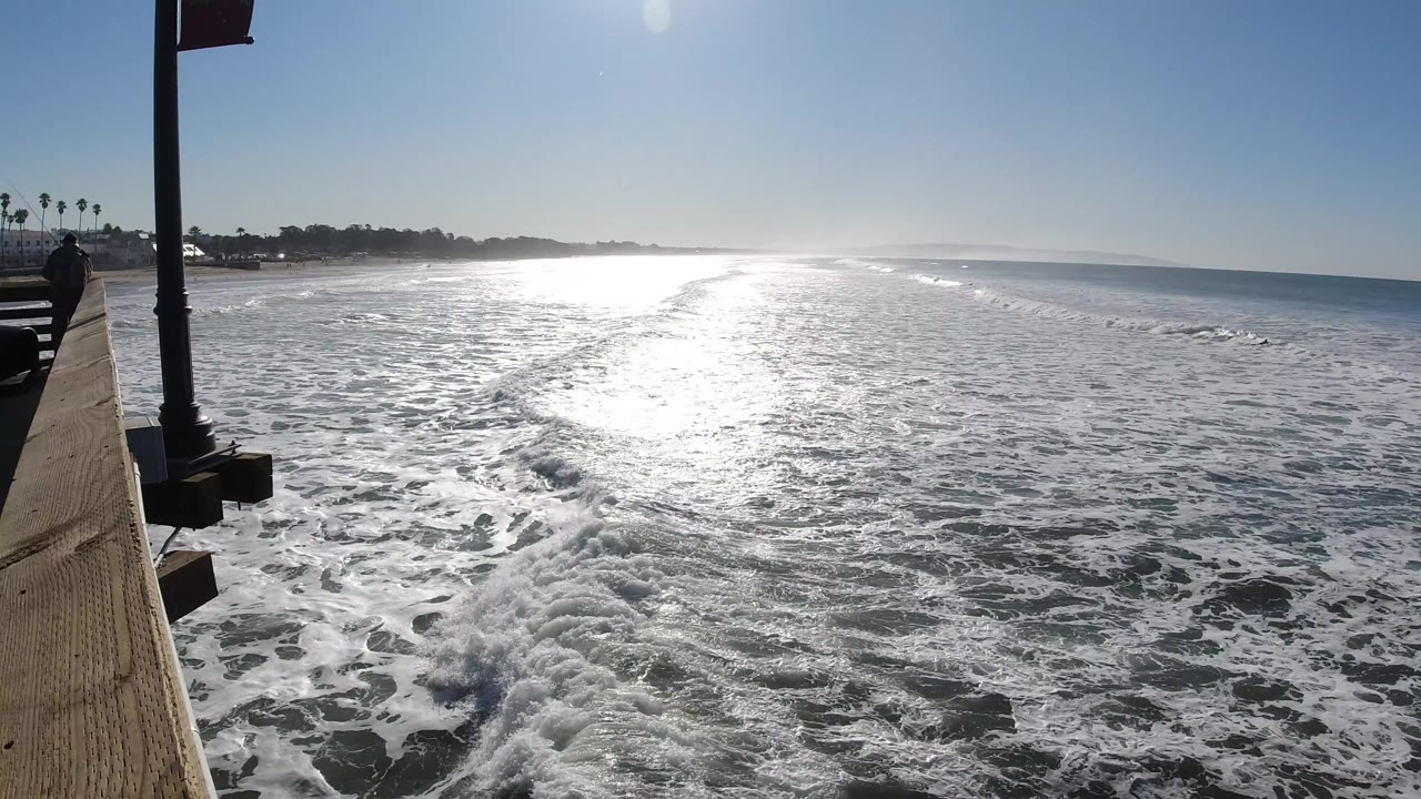 A slightly later Pismo Beach view shows the white water pattern shifting under the sun path, with the pier edge still framing the left side and no visible people.
