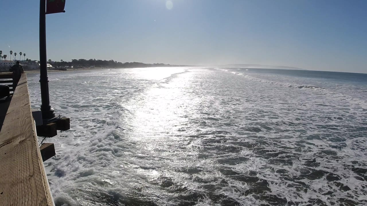 Pismo Beach waves extend toward a low horizon while bright glare runs through the center of the water and the left edge of the pier remains visible with no visible people.