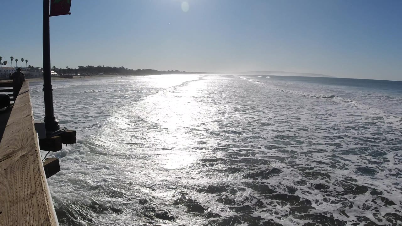 Sunlit ocean foam and small waves beside a pier railing.