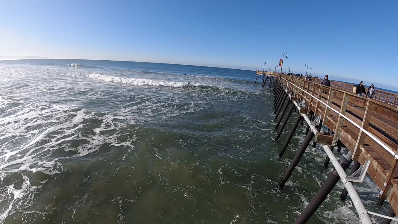 At 0:02, the wooden pier occupies the right edge while textured ocean water and breaking white surf fill the left under a cloudless sky.