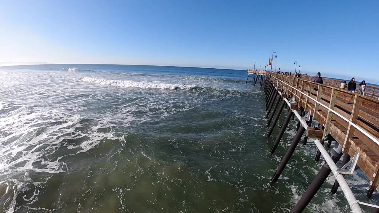 Pismo Beach surf spreads toward shore beside a wooden pier, with bright sun glare at left and small people visible along the right-side walkway.