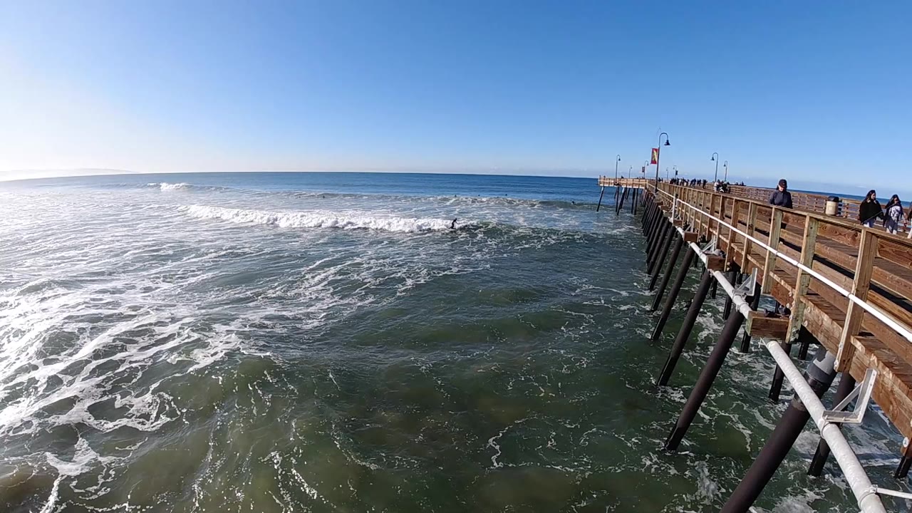 Pismo Beach, California surf spreads across the left side of the frame while a wooden pier with several people along the railing extends from the right.
