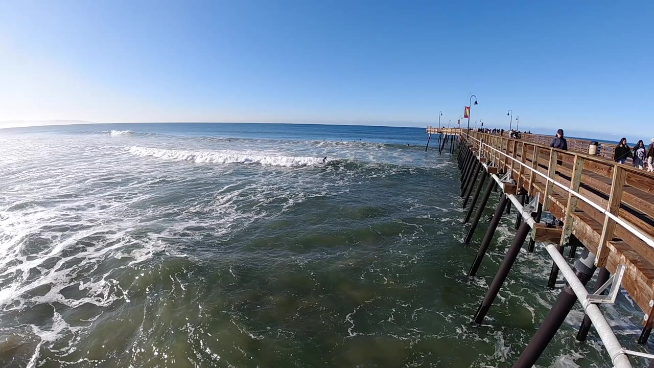 A wave breaks farther across the left half of the Pismo Beach frame as the wooden pier and its dark support pilings angle into the scene from the right.