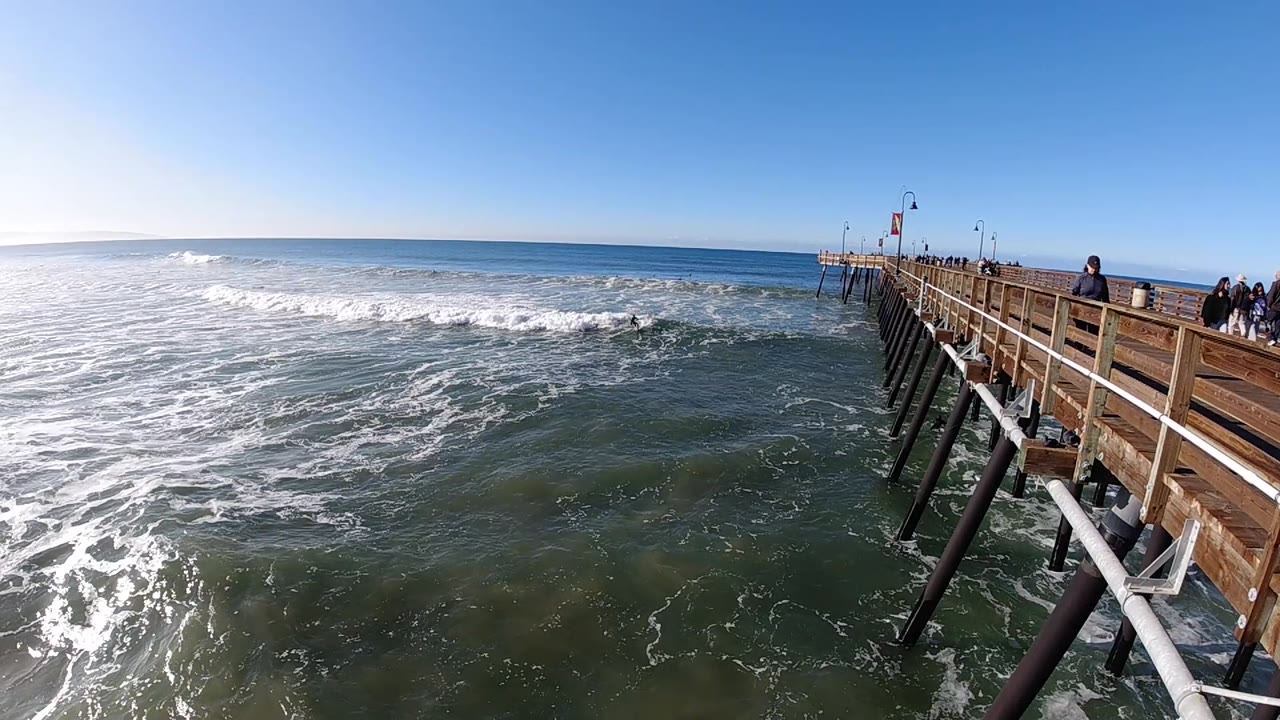 At 0:02, white foam spreads across the center-left water while the Pismo Beach pier and its angled support posts frame the right edge with people still visible above.