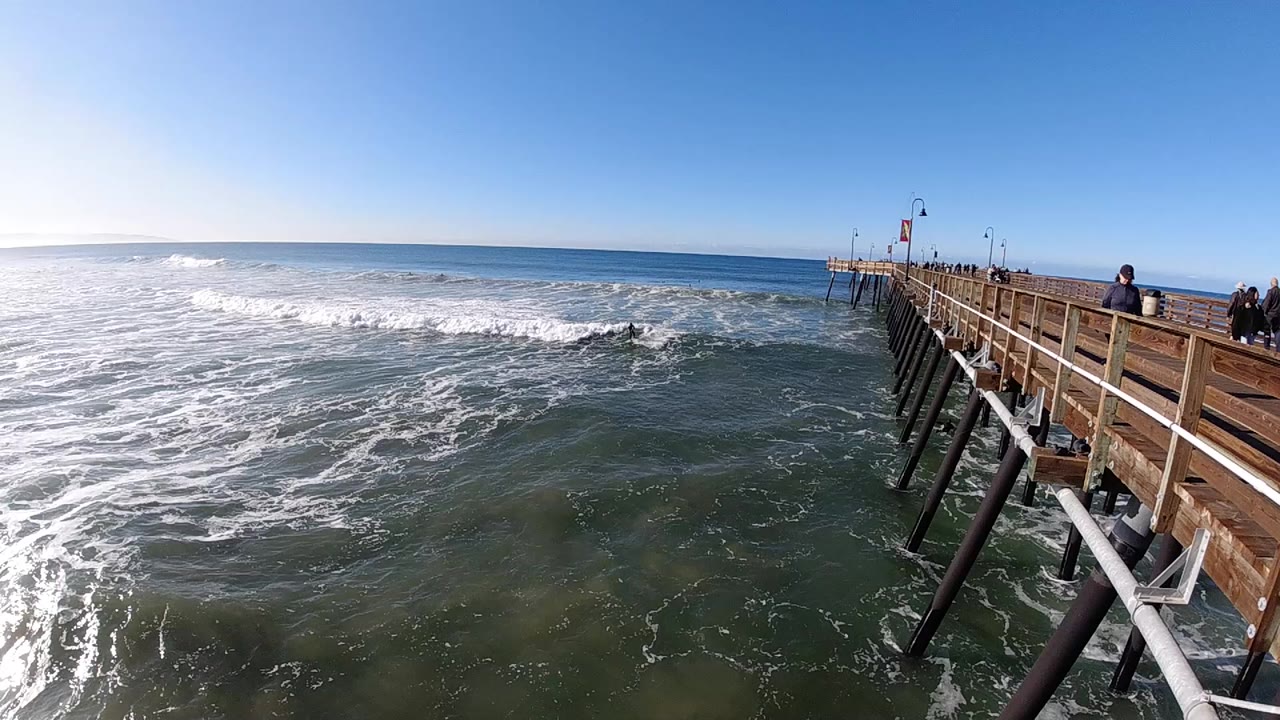 Pismo Beach ocean view with white surf spreading across the water and a wooden pier angling in from the right under clear blue sky.