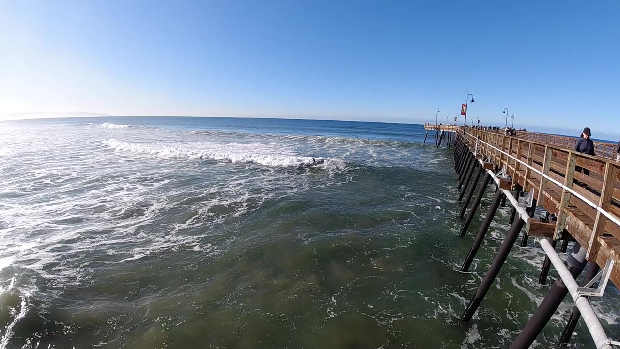 Wide view at Pismo Beach, California, with the wooden pier entering from the right, white surf spreading across the water, bright sun glare on the left, and small people visible on the pier.