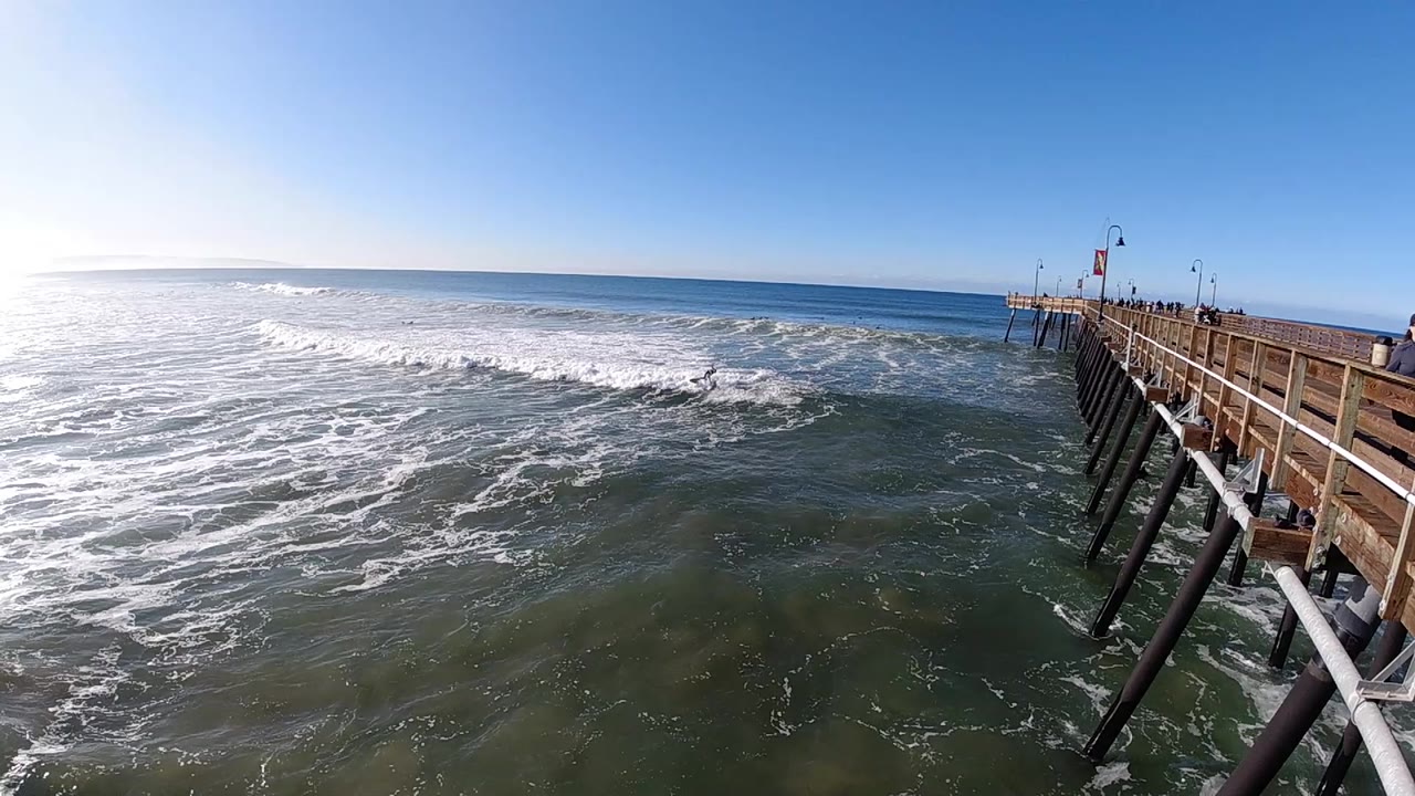 Pismo Beach surf breaks across the left side of the frame while the pier railings and pilings line the right side, with no recognizable people visible.