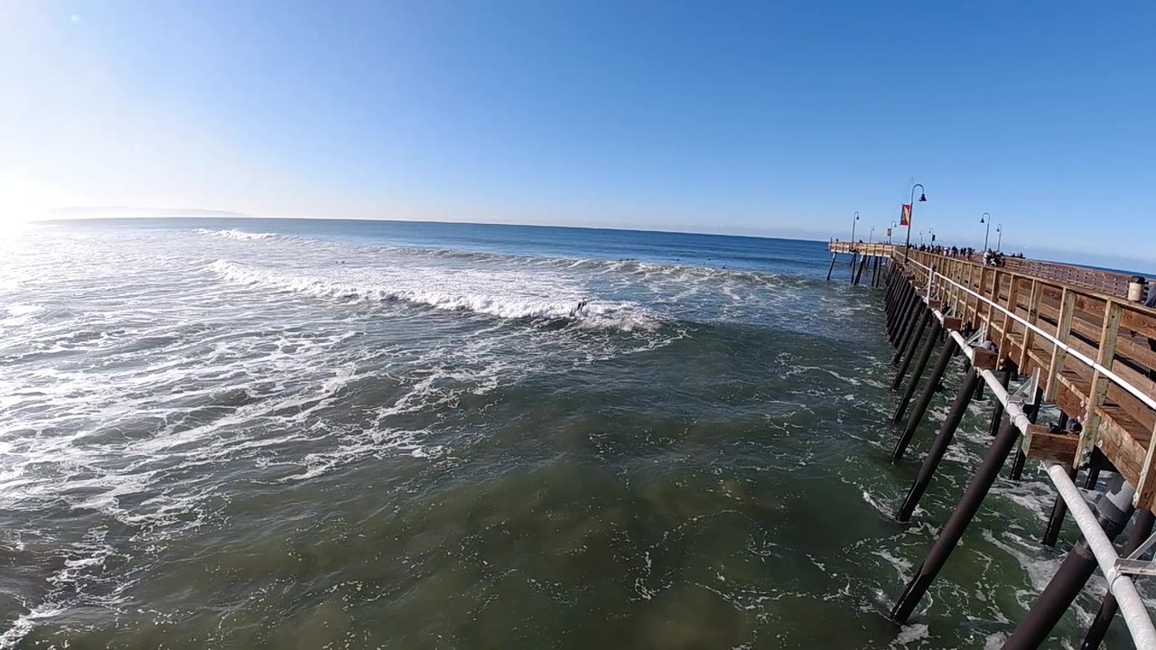 A breaking wave curls toward the pier pilings at Pismo Beach, with sunlit foam, green-blue water, and no recognizable people visible.