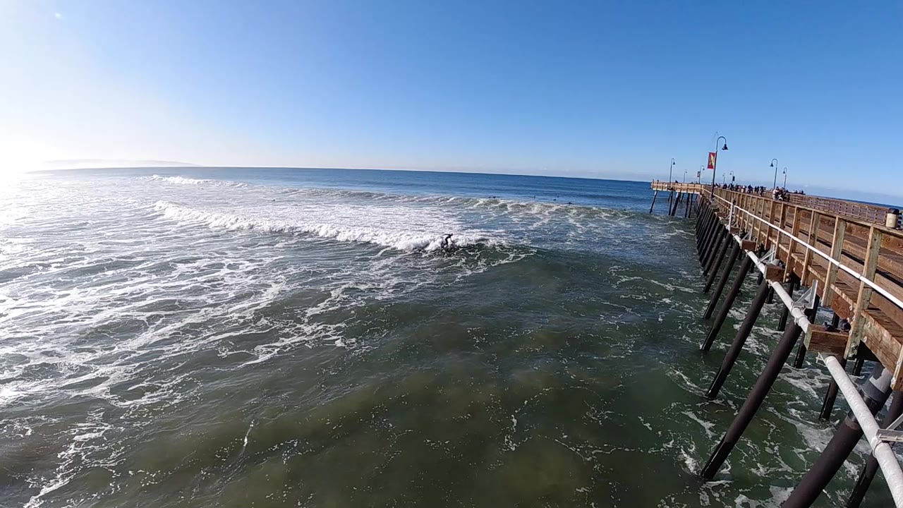 At 0:00, large white surf spreads across the ocean beside the wooden pier in Pismo Beach, California, with blue sky and a bright horizon in the background.