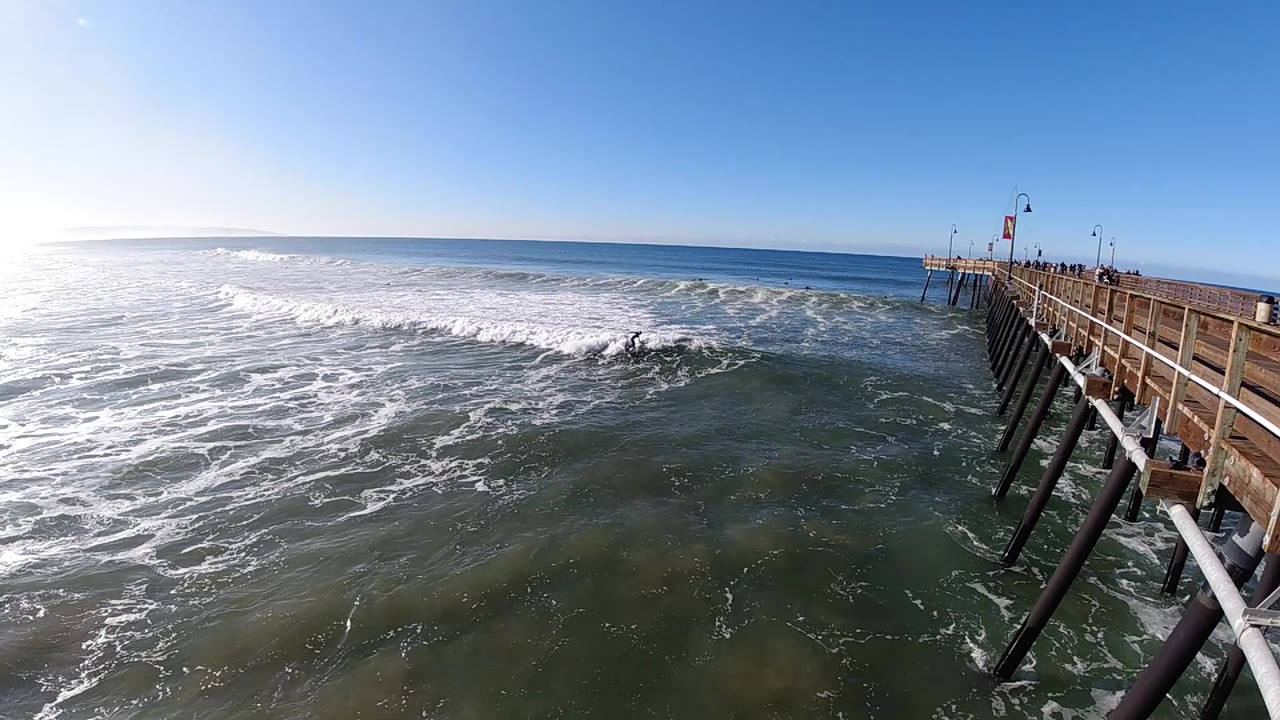 At 0:01, the wave line sits slightly farther across the midground as whitewater rolls toward the pier pilings at the right side of the Pismo Beach frame.