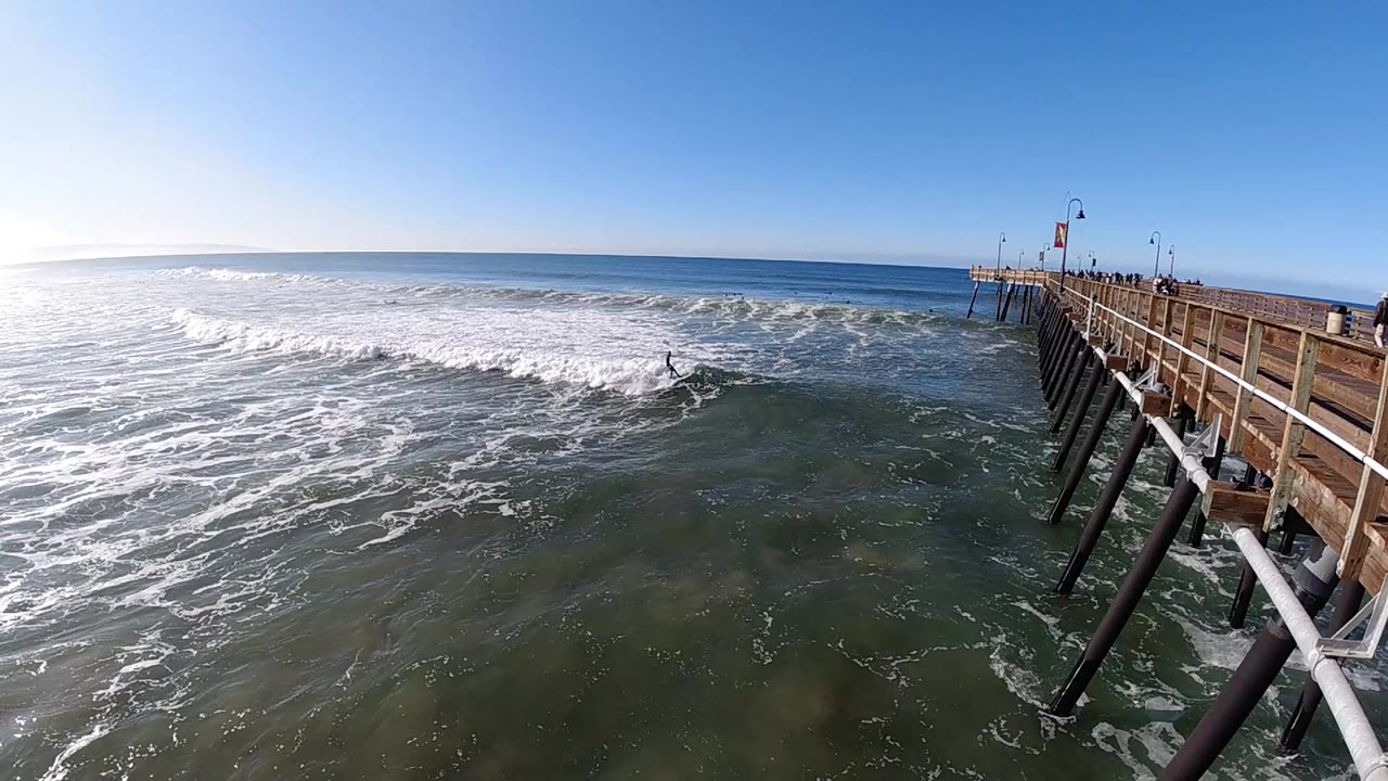At 0:00 in Pismo Beach, a surfer appears in churning water left of a wooden pier, with bright sky and the ocean horizon beyond.