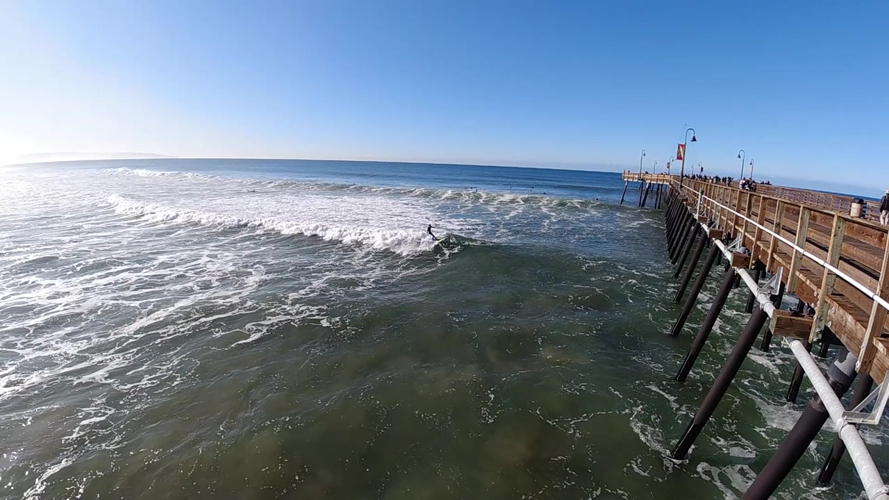 At 0:02 in Pismo Beach, the surfer is centered near the break line as whitewater rolls toward the pier pilings on the right.