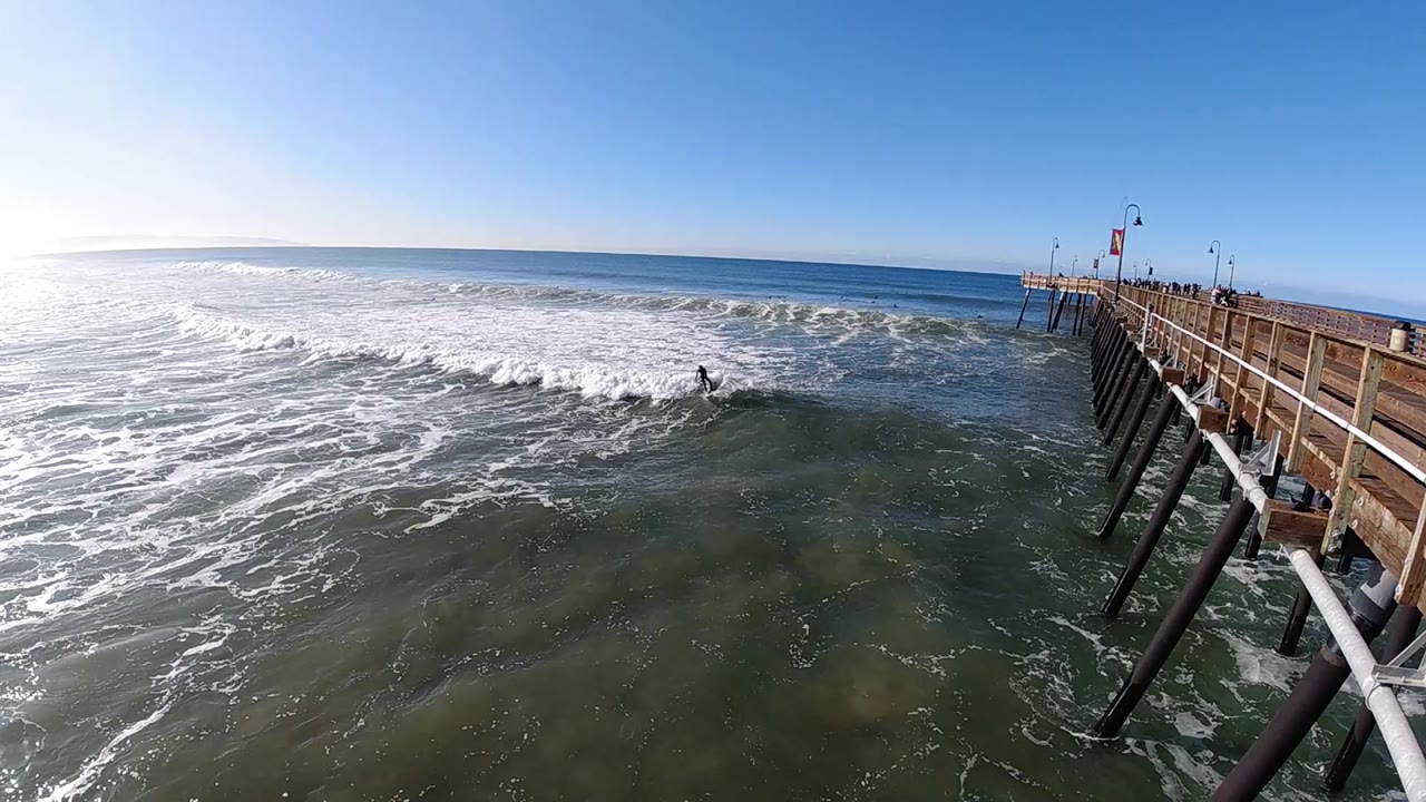 At Pismo Beach, California, a surfer rides through whitewater near the pier while the sun reflects across the ocean and small people stand along the pier rail.