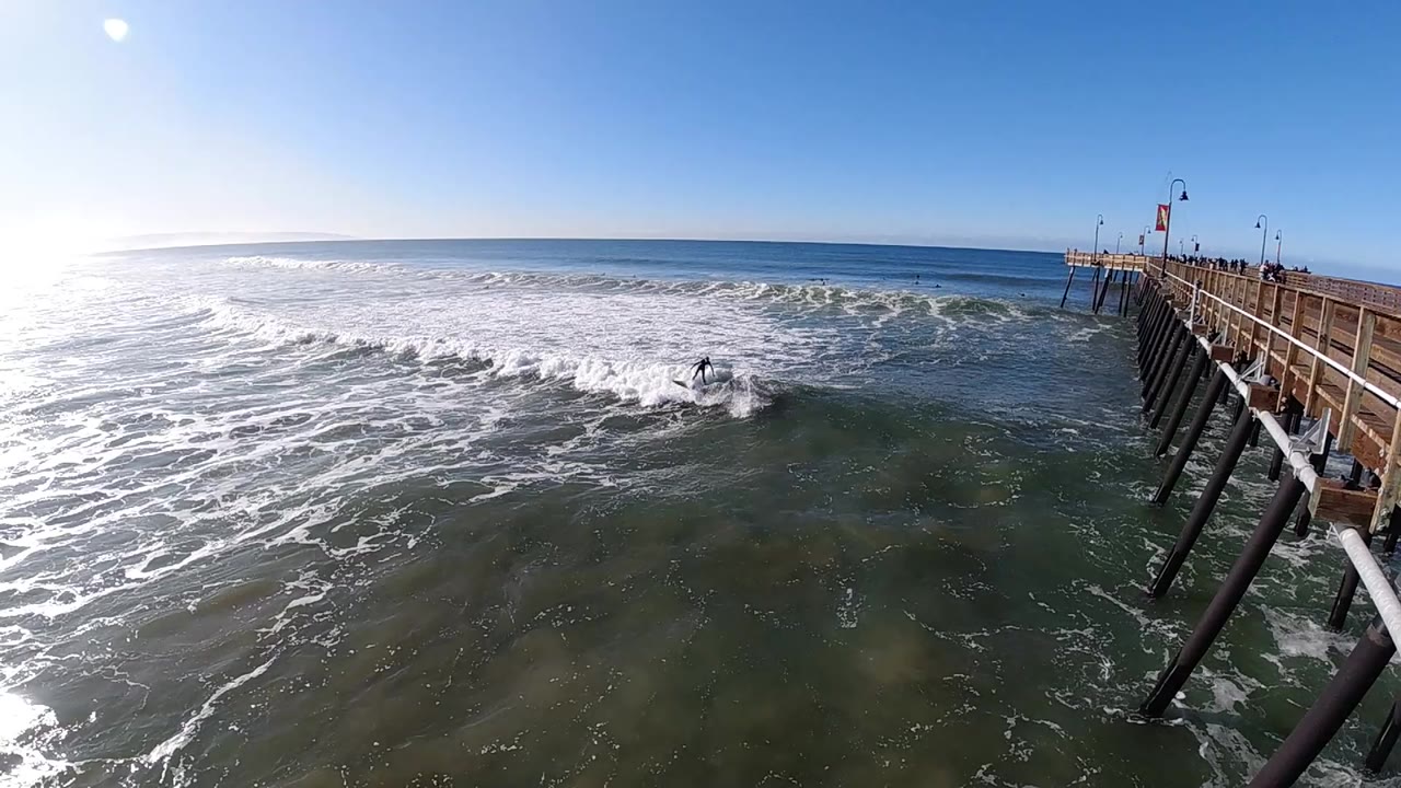 At 0:00 in Pismo Beach, California, whitewater spreads across the ocean beside a wooden pier with dark pilings and bright sun glare at the left edge.
