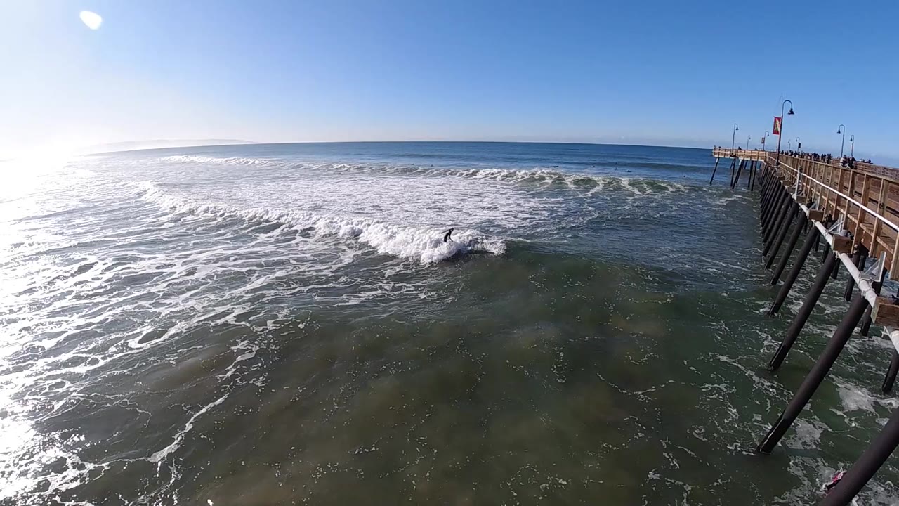 A high wide view at Pismo Beach shows a white wave spreading across green-blue water, bright sun glare on the left, and the pier pilings along the right edge.