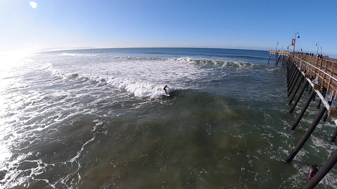 At Pismo Beach, California, the surfer remains in the midground below the pier as a band of white water rolls in behind them and sun glare brightens the left side.