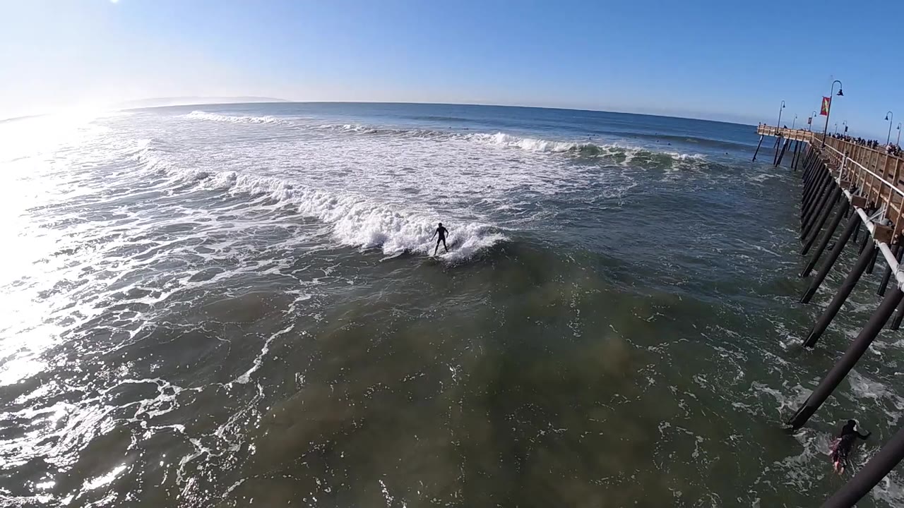 The surfer remains centered in the breaking wave at Pismo Beach while the pier lines the right edge and sunlit foam fills the left side of the frame.