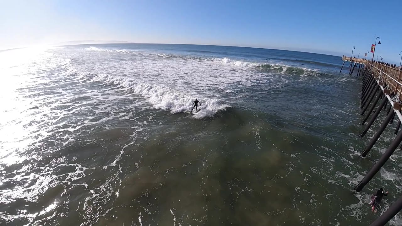 A surfer moves through a breaking wave closer to the pier at Pismo Beach, with the open horizon beyond and bright reflected light across the left water.