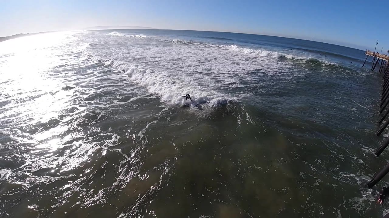 At 0:03, foamy surf rolls diagonally across the green-gray water at Pismo Beach with the quay structure still visible at the right, with no visible people.