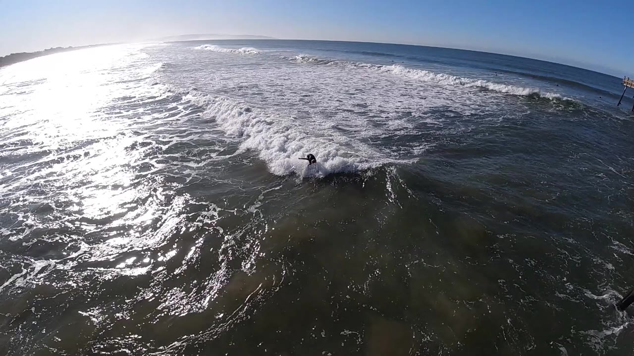 At Pismo Beach, California, a small surfer rides near the lower center of a wide aerial ocean frame as sun glare spreads across the left side and whitewater breaks behind.
