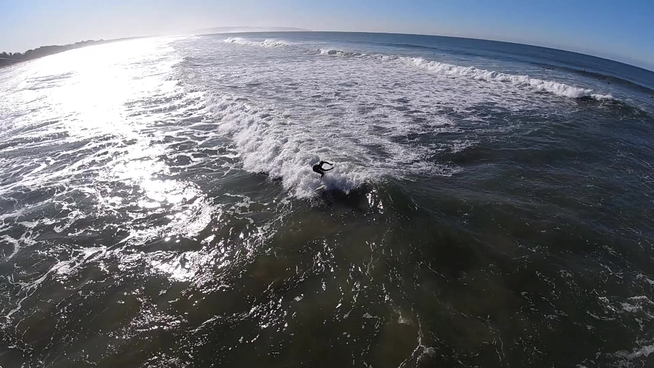 A surfer in a dark wetsuit rides whitewater near Pismo Beach, California, with intense sun reflection across the left side of the ocean and a tilted horizon above.