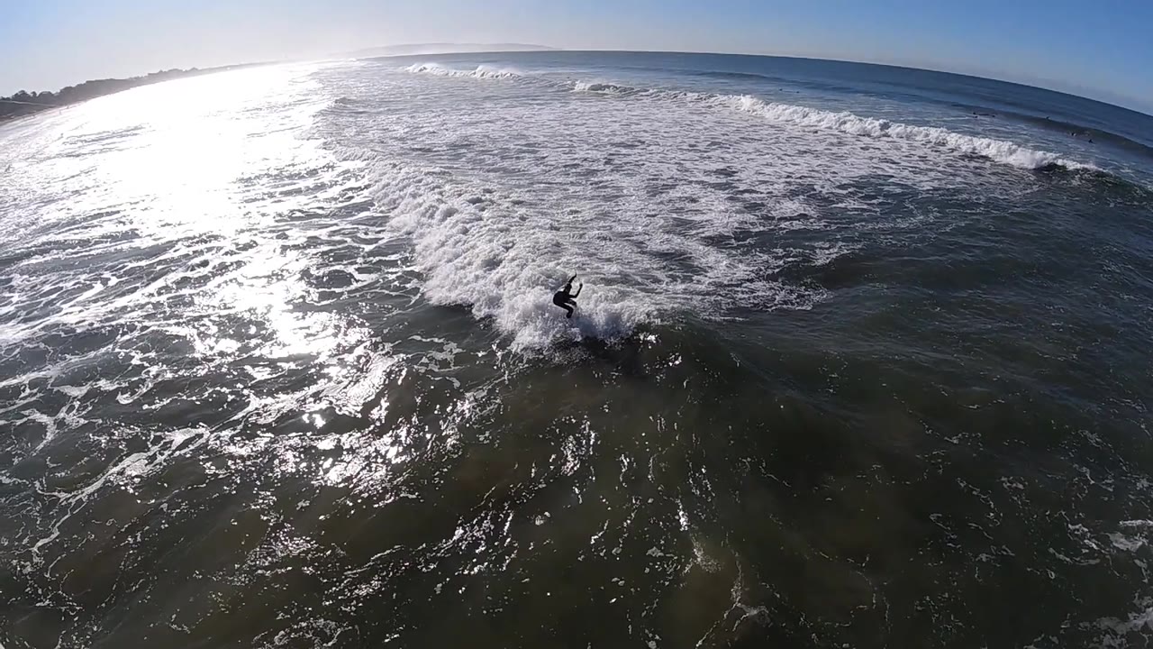 A lone surfer rides near the lower center of the frame at Pismo Beach, California, with sunlit chop to the left and a band of breaking surf stretching across the background.