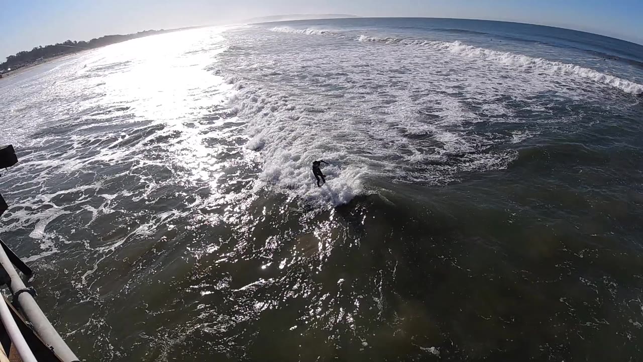 A distant man rides across foamy sunlit surf at Pismo Beach, California, with a pier-side railing cutting into the lower edge and glare brightening the left side of the water.
