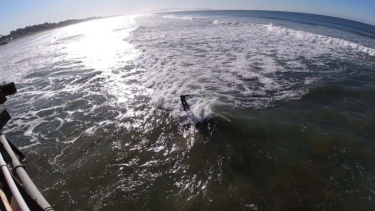 The man appears farther right in the lower surf at Pismo Beach as textured whitecaps fill the midground and the coastline remains faint along the upper left horizon.