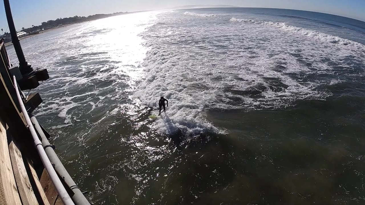 The surfer remains upright in the lower part of the Pismo Beach frame, with pier structure along the left side and a textured white wave line filling the midground.
