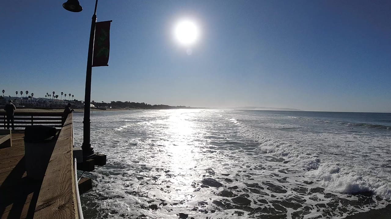 Bright sun reflects across foamy waves in Pismo Beach, California, with a pier-side railing and lamp post at the left and no visible people.