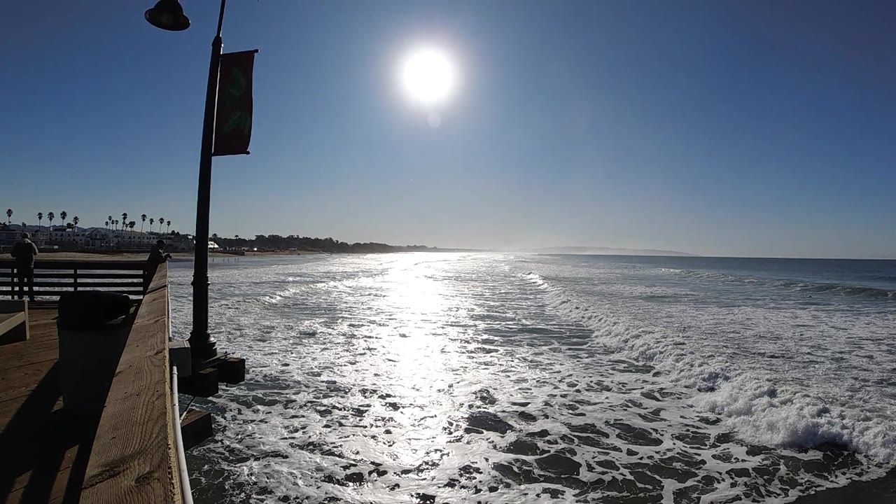 Foam spreads across the near water in Pismo Beach, California, while the pier railing and lamp post remain at left and no visible people appear.