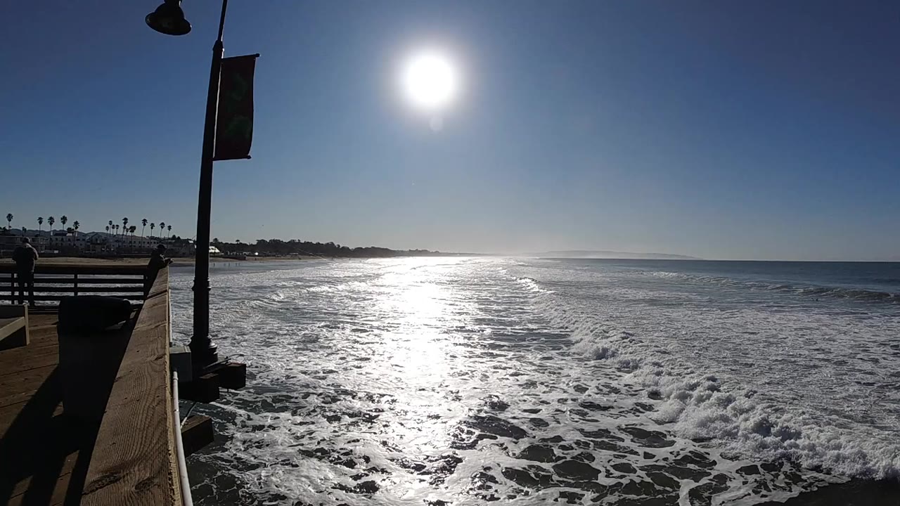 At 0:00 in Pismo Beach, bright sun reflects across foamy ocean water beside a platform railing and lamp post, with no clearly visible people.