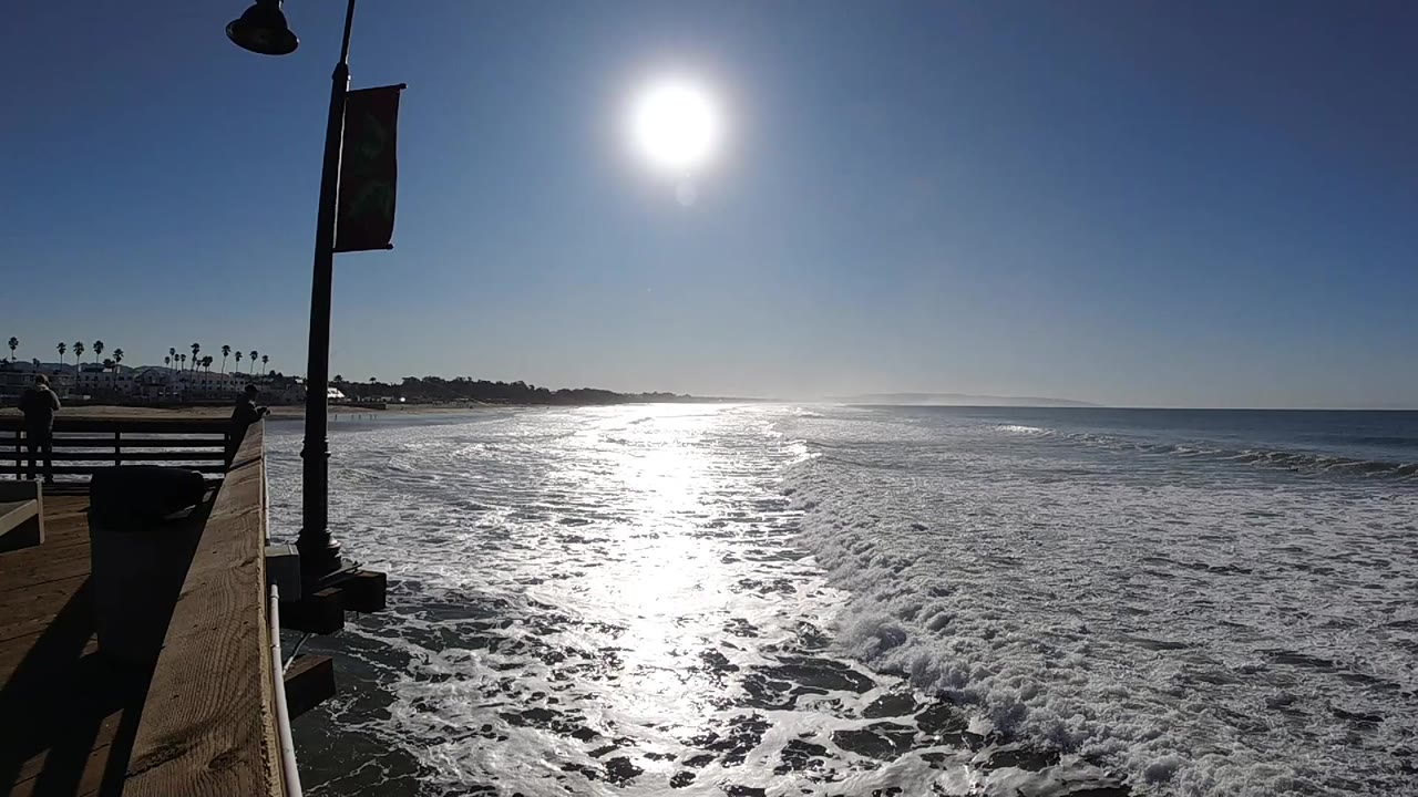 Pismo Beach surf fills the frame under a high sun, with the pier railing and lamp post on the left and no visible people.