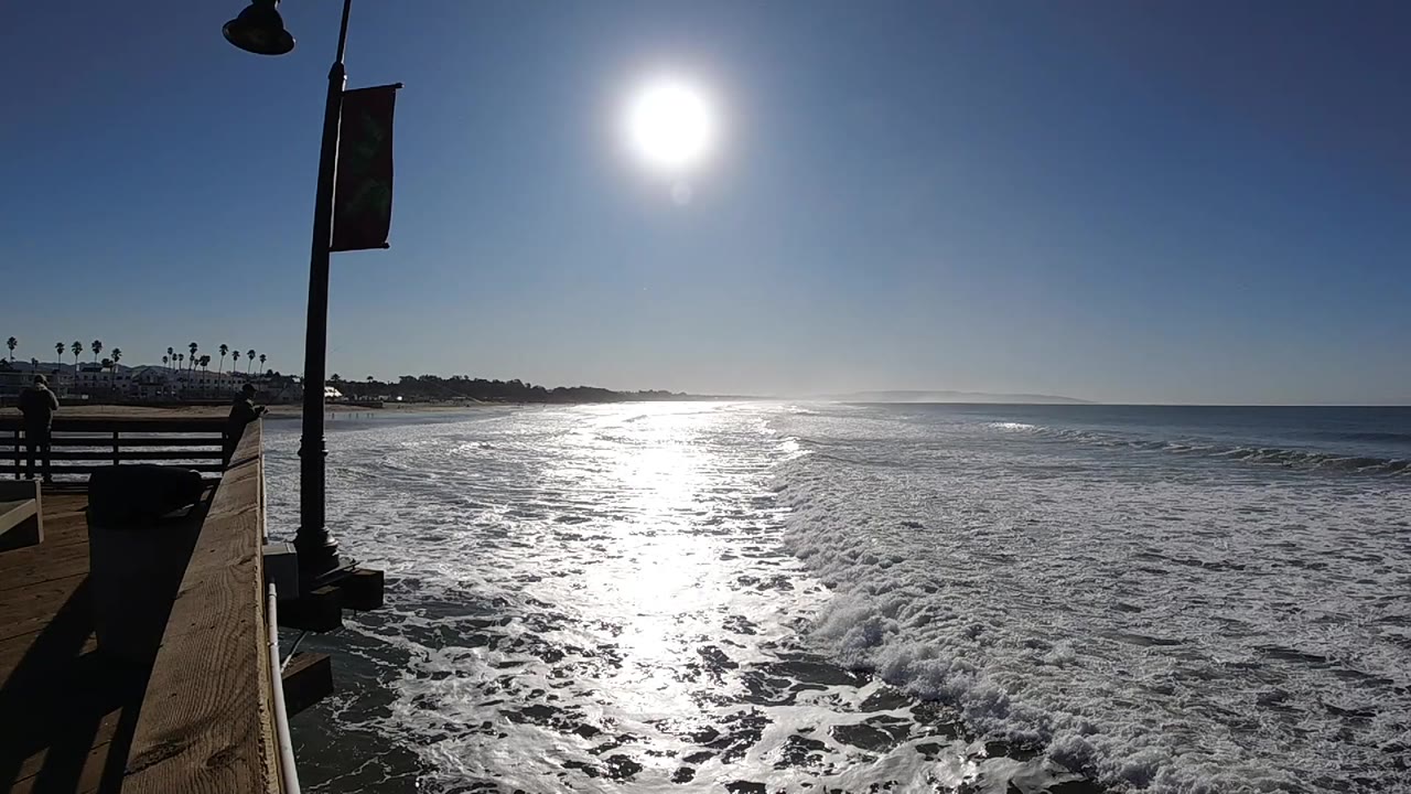 Pismo Beach waves break closer to the pier while the sun glare spreads over the water, with shoreline and palms visible to the left and no visible people.
