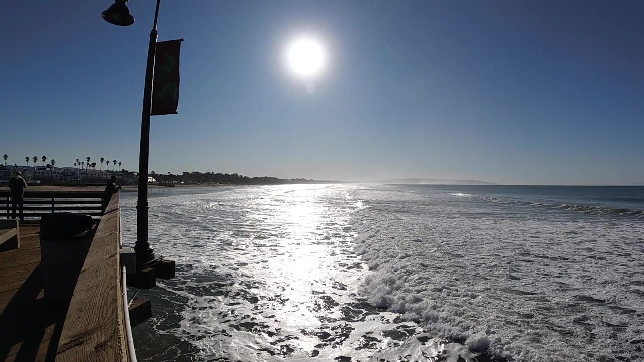 Pismo Beach ocean surface appears flatter near the horizon while white foam spreads below the pier, with a lamp post silhouetted at left and no visible people.