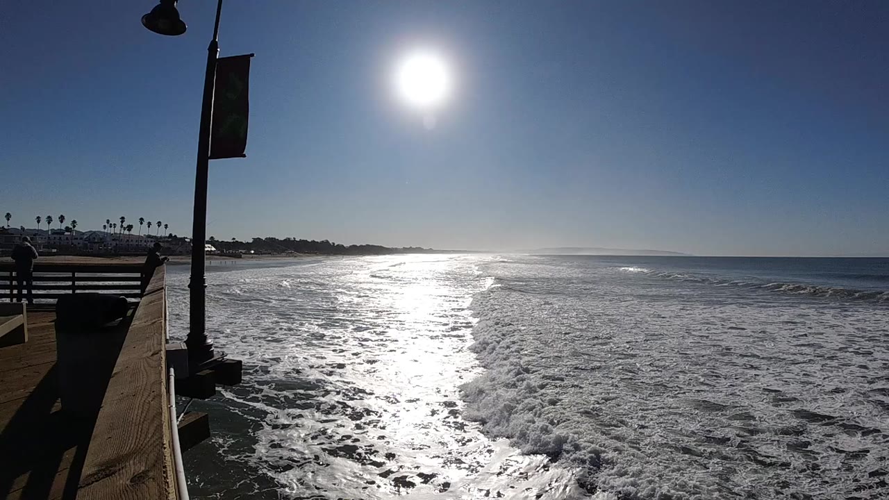 Opening frame of Pismo Beach surf under a high bright sun, with the pier railing and lamp post at left and no visible people.