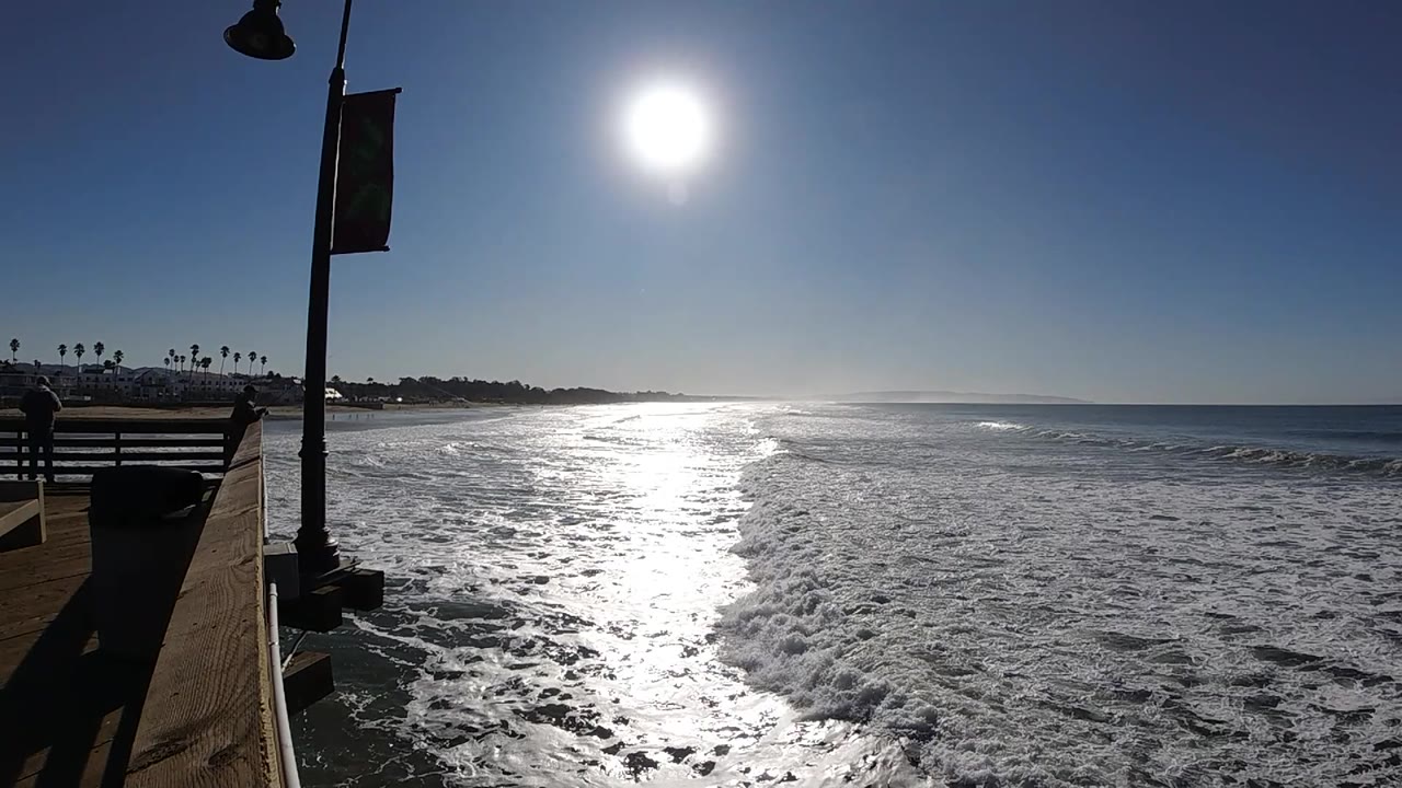 A broad sheet of whitewater rolls toward shore at Pismo Beach, with the sun high above and the pier structure still visible at left, no visible people.