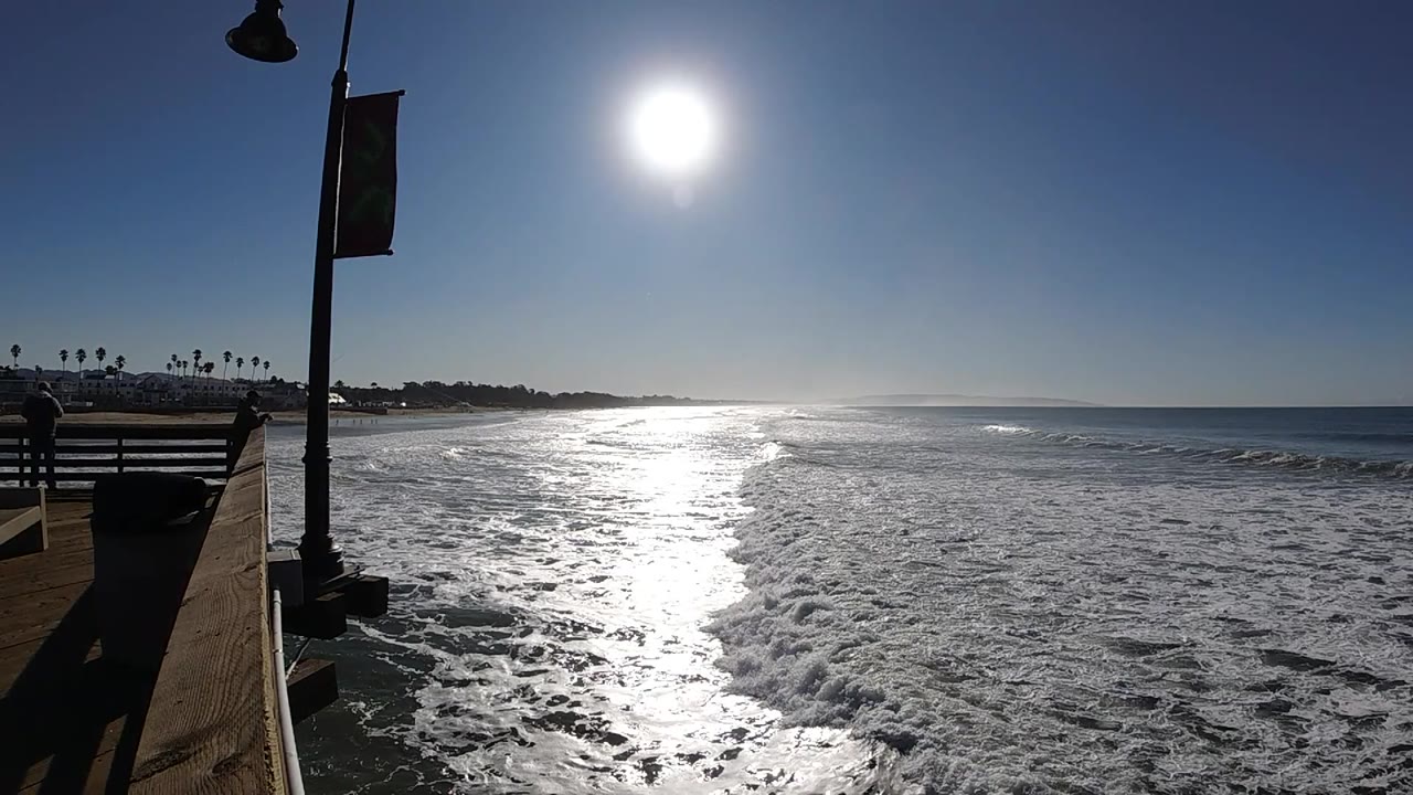 Pismo Beach surf reflects the overhead sun while the shaded pier edge, railing, lamp post, and banner create a dark foreground border, with no visible people.