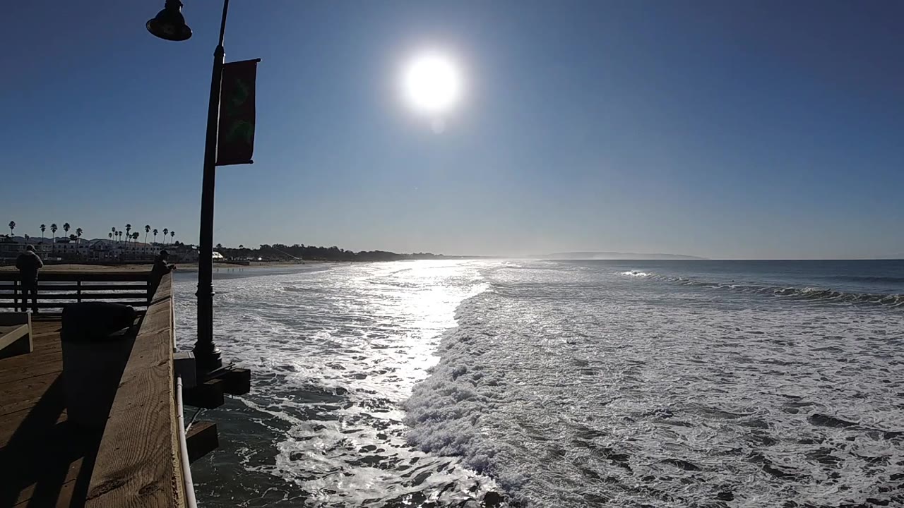 Sunlit Pismo Beach waves break in a diagonal line toward the lower right, with the promenade railing and lamp post still framing the left side and no visible people.