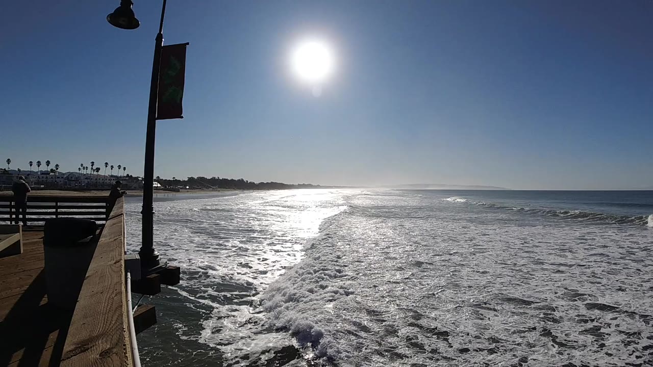 Pismo Beach ocean surf rolls toward shore beside a dark pier railing and lamp post, with bright sun glare on the water and no visible people.