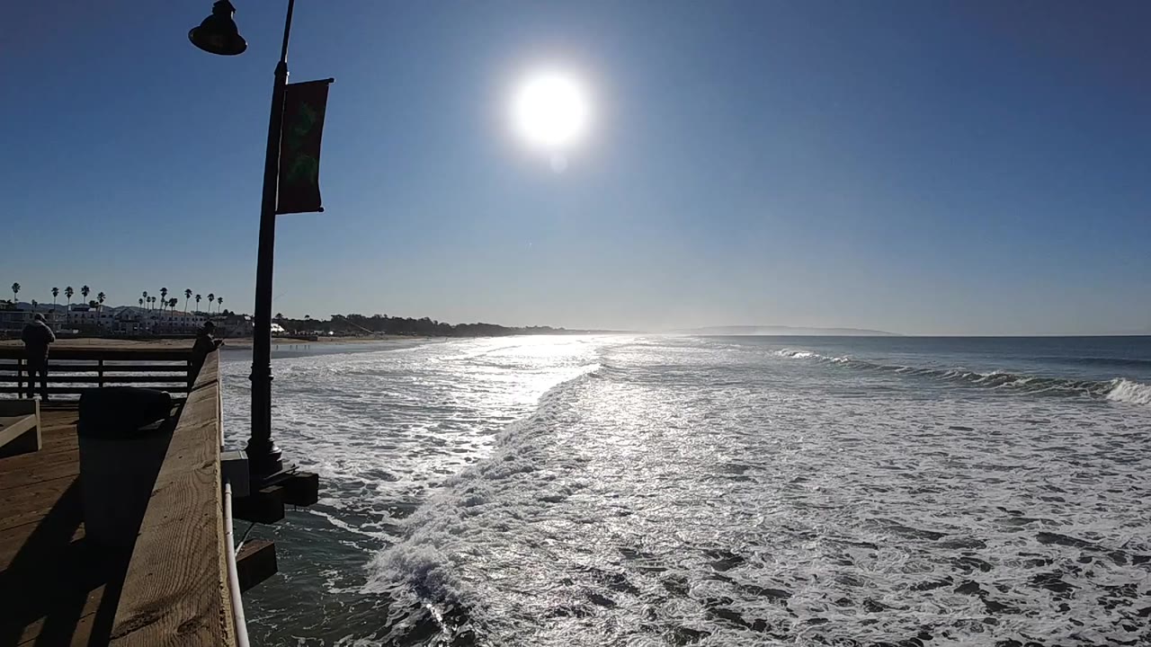 Pismo Beach surf spreads across the frame under a high sun, with a pier railing and lamp post at left and no visible people.