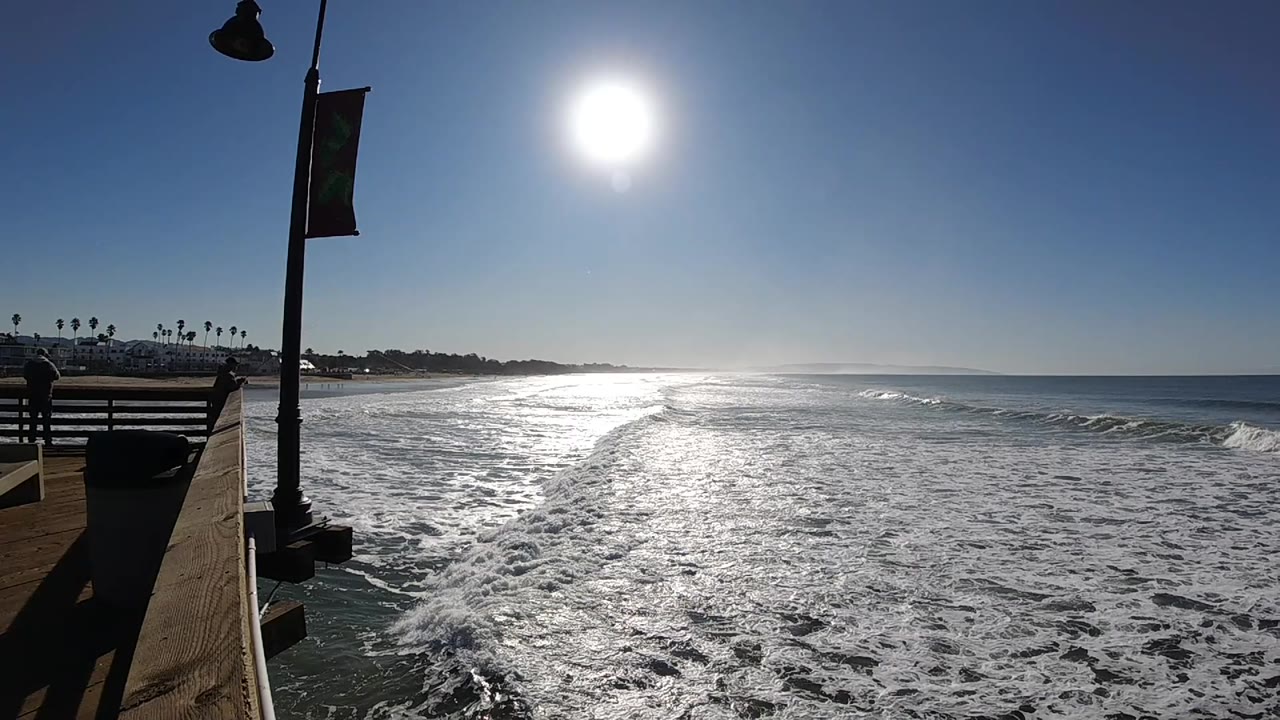 At 0:02, the Pismo Beach shoreline remains distant while the wave line sits farther right, with bright glare across the water and no visible people.