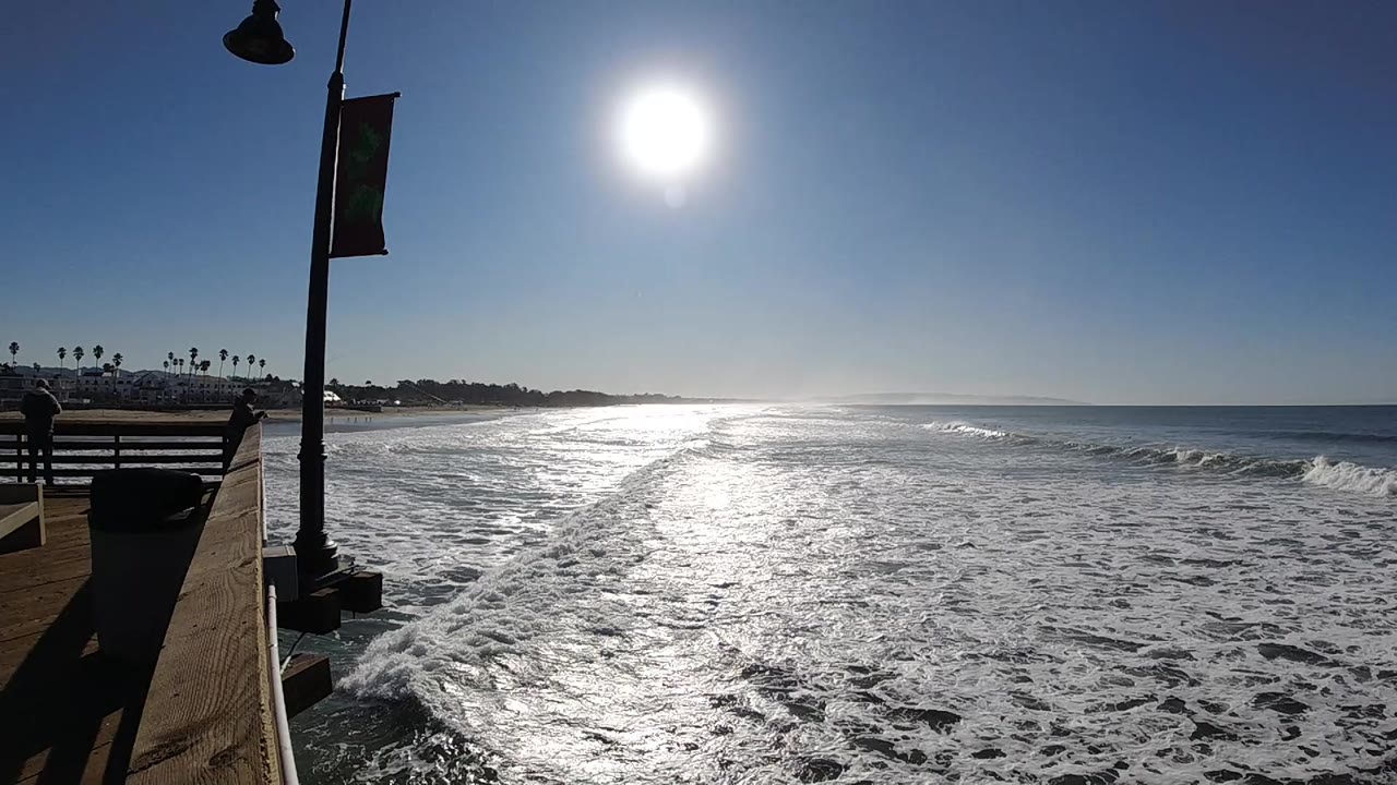 Opening frame of Pismo Beach surf with bright sun glare on foamy water, a pier-side railing and lamppost at left, and no visible people.