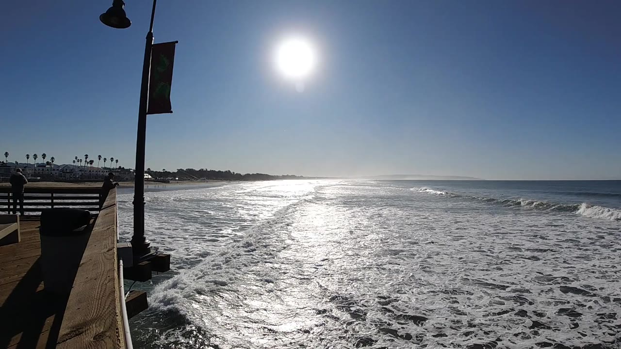 Mid-clip Pismo Beach frame with a small breaking wave to the right, glare across the water, foreground lamppost, and no visible people.