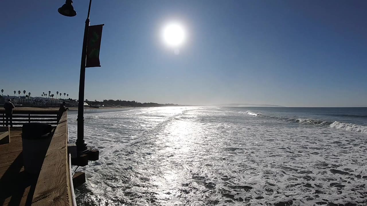Sunlight streaks across the water at Pismo Beach, California, while low waves break near shore and the left edge shows railing and lamp posts with no visible people.