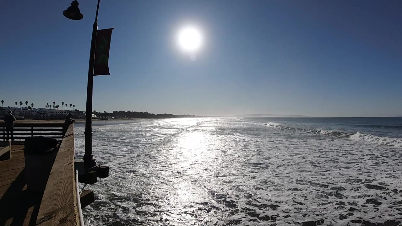 At Pismo Beach, bright sun reflects across foamy surf beside a boardwalk railing, lamp post, banner, and distant palm trees, with no visible people.