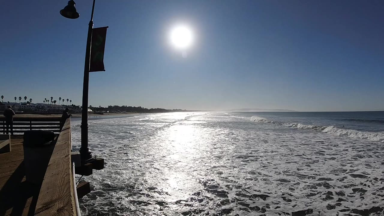 Foamy waves roll under bright sun with pier railing at left.