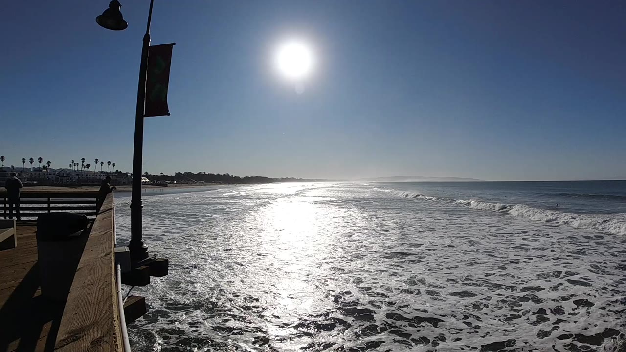 Bright sun reflects off foamy waves and wet sand at Pismo Beach, California, with a lamp post and pier edge at left and no visible people.