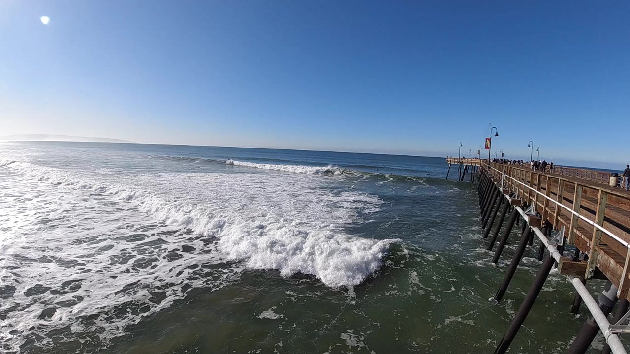 At 0:00 in Pismo Beach, white surf breaks across the foreground beside a wooden pier on the right, with a bright sun flare at upper left and no identifiable people visible.